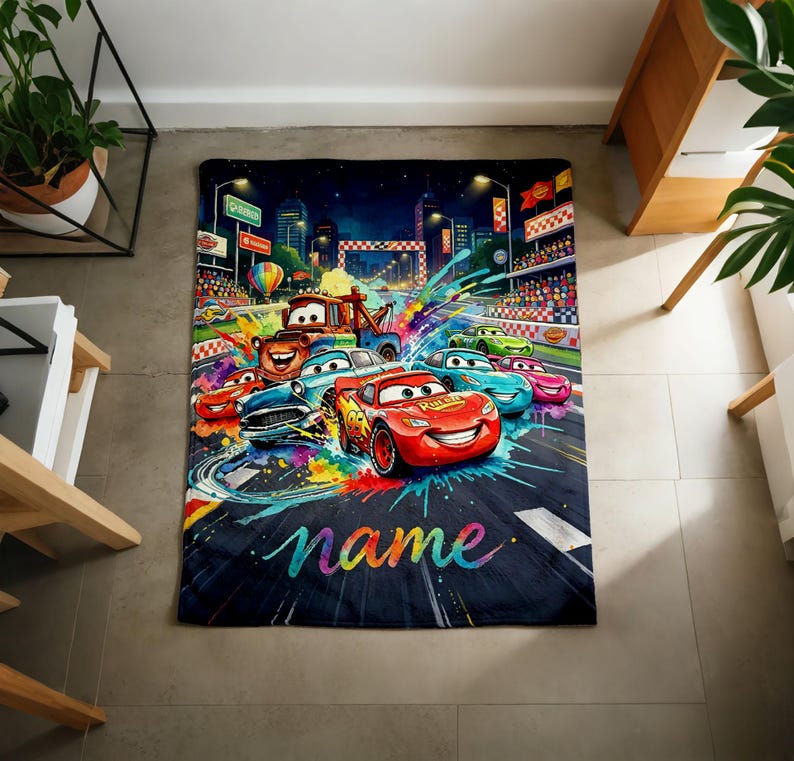 Puede incluir: Una colorida manta con coches de dibujos animados corriendo en una pista. El dise&ntilde;o incluye un fondo de paisaje urbano, una l&iacute;nea de meta y la palabra "name" en una fuente de arco&iacute;ris. La manta es una vibrante mezcla de colores.