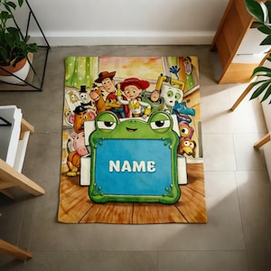 May include: A rectangular fleece blanket featuring a colorful Toy Story character print. In the center, a large green frog-shaped frame with a blue rectangular insert displays the white text NAME. The background shows various animated characters.