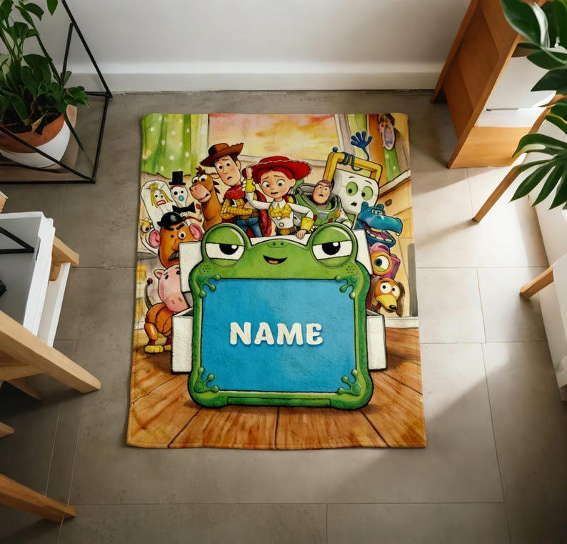May include: A rectangular fleece blanket featuring a colorful Toy Story character print. In the center, a large green frog-shaped frame with a blue rectangular insert displays the white text NAME. The background shows various animated characters.