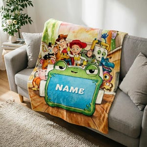 May include: A soft fleece throw blanket featuring a colorful Toy Story character print with Woody, Jessie, Buzz Lightyear, and others. In the foreground, a large green frog-shaped frame with a blue center displays the white text NAME.