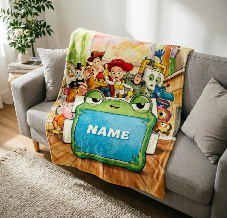 May include: A soft fleece throw blanket featuring a colorful Toy Story character print with Woody, Jessie, Buzz Lightyear, and others. In the foreground, a large green frog-shaped frame with a blue center displays the white text NAME.