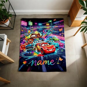 Puede incluir: Una colorida manta con tem&aacute;tica de Cars que presenta a Rayo McQueen y otros personajes compitiendo en una pista. El dise&ntilde;o incluye colores ne&oacute;n vibrantes, un fondo azul oscuro y la palabra "name" en cursiva.