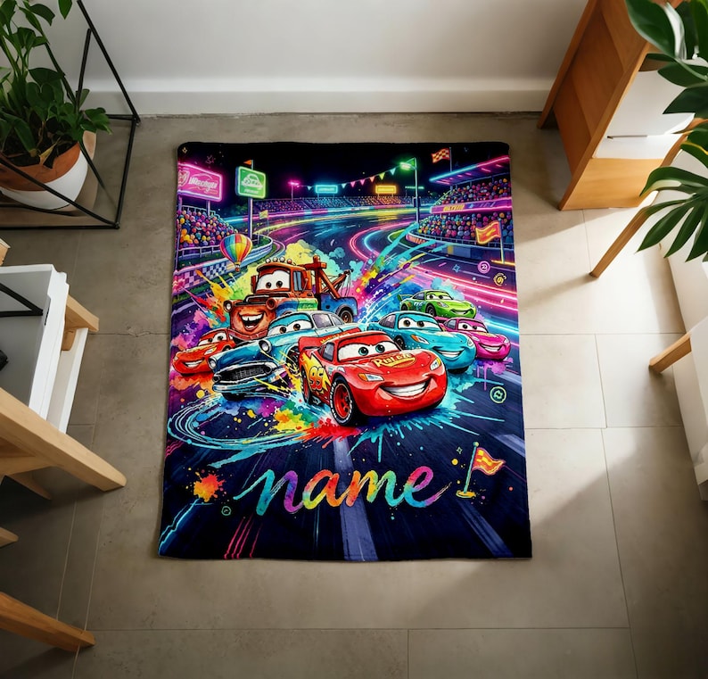 Puede incluir: Una colorida manta con tem&aacute;tica de Cars que presenta a Rayo McQueen y otros personajes compitiendo en una pista. El dise&ntilde;o incluye colores ne&oacute;n vibrantes, un fondo azul oscuro y la palabra "name" en cursiva.