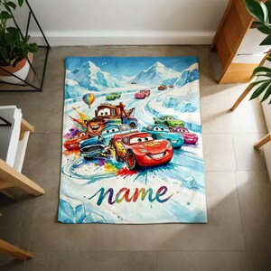 Puede incluir: Una manta rectangular con una colorida ilustraci&oacute;n de coches de carreras de dibujos animados en una pista nevada. El dise&ntilde;o incluye un coche de carreras rojo con el texto "name" en una fuente arco&iacute;ris. El fondo muestra monta&ntilde;as cubiertas de nieve y un cielo azul.