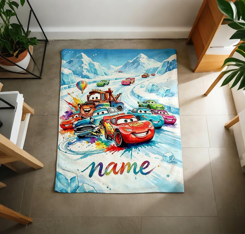 Puede incluir: Una manta rectangular con una colorida ilustraci&oacute;n de coches de carreras de dibujos animados en una pista nevada. El dise&ntilde;o incluye un coche de carreras rojo con el texto "name" en una fuente arco&iacute;ris. El fondo muestra monta&ntilde;as cubiertas de nieve y un cielo azul.