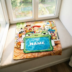 May include: A rectangular fleece blanket featuring a colorful print of various animated characters from the Toy Story franchise. In the foreground, a large green frog character holds a blue rectangular sign with the white text NAME.