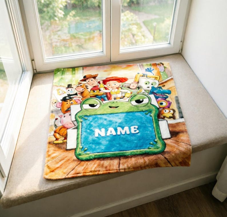May include: A rectangular fleece blanket featuring a colorful print of various animated characters from the Toy Story franchise. In the foreground, a large green frog character holds a blue rectangular sign with the white text NAME.