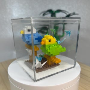 Op de afbeelding: Een heldere acryl display case bevat twee gepixelde vogelsculpturen, een blauwe en een groene, die op bruine platforms zitten. Origami-kranen en blauwe vlinders voegen details toe. De case staat op een witte draaiende standaard.