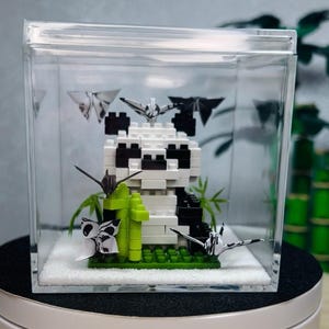 Op de afbeelding: Een pandabeerfiguur gemaakt van witte en zwarte blokken, met groene bamboe en origami-kranen, in een doorzichtige acrylbox. De doos staat op een wit oppervlak.