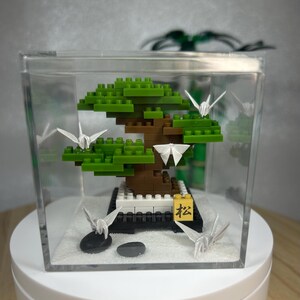 Op de afbeelding: Een miniatuur bonsai-boom gemaakt van groene en bruine blokken, geplaatst in een heldere acryl behuizing. Witte origami-kranen en kleine zwarte stenen zijn rondom de boom geplaatst. Een klein geel blok met Japanse karakters is ook aanwezig.