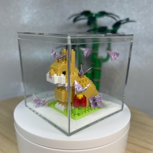 Könnte beinhalten: Eine gelbe, aus Ziegeln gebaute Kaninchenfigur mit weißer Nase und Ohren in einer transparenten Vitrine. Origami-Kraniche und -Blumen umgeben das Kaninchen, mit einer grünen Basis und einem roten Block. Bambus ist im Hintergrund zu sehen.
