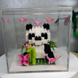 Könnte beinhalten: Eine Pandabären-Skulptur aus weißen und schwarzen Blöcken in einer transparenten Vitrine. Rosa Origami-Kraniche und -Blumen sowie grüner Bambus schmücken die Szene. Der Sockel ist mit weißem Glitzer versehen.