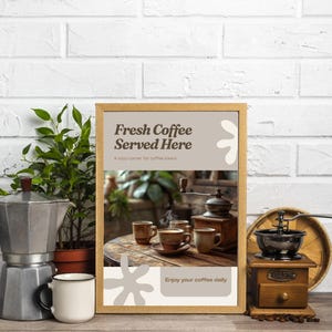 Puede incluir: Un letrero enmarcado con el texto "Fresh Coffee Served Here" y una imagen temática de café. Junto al letrero hay una cafetera exprés plateada, una taza blanca y una pequeña planta en maceta. También se ve un molinillo de café de madera.