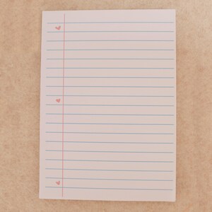 Pode incluir: Um bloco de notas branco com linhas horizontais azuis e uma margem vertical rosa. Pequenos corações rosa são impressos ao longo da margem. O bloco de notas está sobre uma superfície castanha clara.