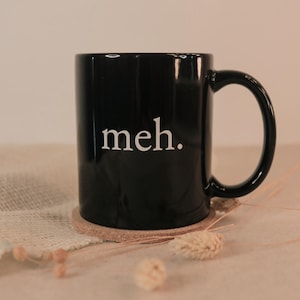 Op de afbeelding: Een zwarte keramische mok met het woord "meh." in witte letters. De mok staat op een ronde, lichtbruine onderzetter. De handgreep is gebogen, effen zwart. De achtergrond is neutraal beige.