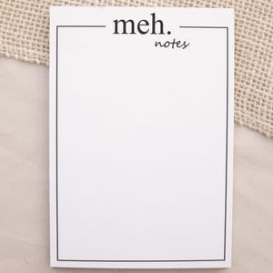 Könnte beinhalten: Weißer rechteckiger Notizblock mit schwarzem Rand. Die Worte "meh. notes" sind in Schwarz gedruckt. Der Notizblock ist leer, mit einem hellbeigen Stoffhintergrund.