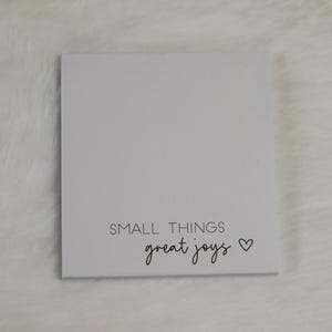 Könnte beinhalten: Ein quadratisches weißes Schild mit dem schwarzen Text SMALL THINGS in einer serifenlosen Schrift, gefolgt von dem Wort great joys in Schreibschrift und einem kleinen schwarzen Herzsymbol rechts neben dem Text.