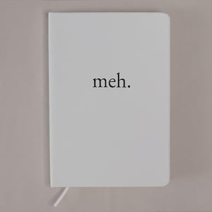 May include: A white hardcover notebook with rounded corners featuring the word meh. printed in a black serif font in the center of the front cover. A thin white ribbon bookmark extends from the bottom edge of the notebook spine.