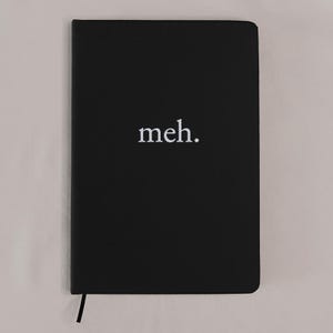 Può includere: Un taccuino rettangolare nero con finitura opaca. La parola "meh." è stampata in bianco, in minuscolo e con un carattere serif al centro della copertina anteriore. Un sottile segnalibro a nastro nero sporge dal bordo inferiore della copertina.