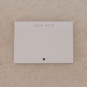 May include: A rectangular, off-white card with the words "LOVE NOTE" printed in a dark gray font at the top. A small black heart is printed at the bottom. The card is on a neutral-toned background.