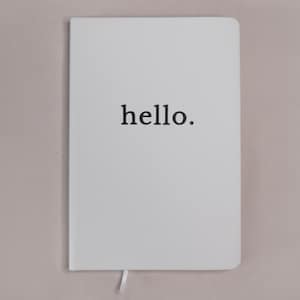 Könnte beinhalten: Ein weißes Notizbuch mit abgerundeten Ecken, auf dessen Vorderseite das Wort hello. in einer schwarzen Serifenschrift zentriert gedruckt ist. Ein kleines weißes Lesebändchen ragt aus der Unterkante des Notizbuchs, das eine glatte Oberfläche hat.