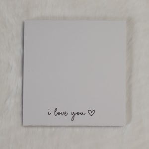 Könnte beinhalten: Eine quadratische weiße Karte mit dem schwarz aufgedruckten Text i love you, gefolgt von einem kleinen, handgezeichneten Herzsymbol. Die Karte hat eine saubere, glatte Oberfläche und ist mittig platziert, mit einer einfachen, dunklen Schrift.