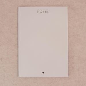 May include: A rectangular, off-white notepad with the word "NOTES" printed at the top. A small black heart is at the bottom. The notepad is on a light pink surface.