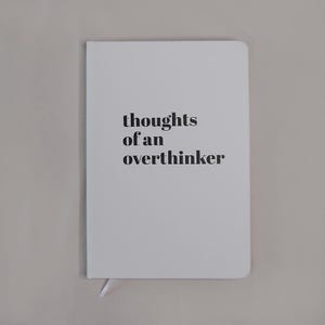 Può includere: Un taccuino bianco con angoli arrotondati che presenta il testo stampato in nero thoughts of an overthinker centrato sulla copertina anteriore. Un piccolo segnalibro a nastro bianco sporge dal bordo inferiore del diario rilegato.