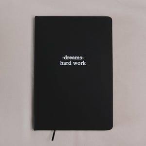 May include: A black rectangular notebook with rounded corners. The front cover features white text that reads "dreams" with a horizontal line striking through it, positioned directly above the word "hard work" in a clean, sans-serif font.