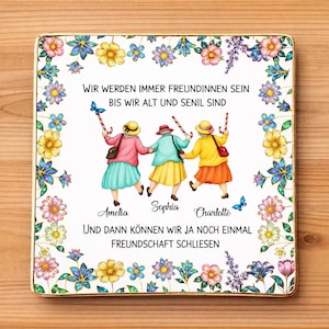 May include: A square ceramic tile featuring a whimsical illustration of three elderly women holding hands, surrounded by a floral border. The tile has text in German, including the names "Amelia," "Sophia," and "Charlotte."