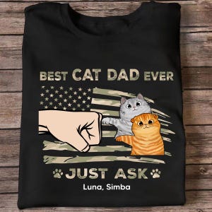Può includere: T-shirt nera con un design grafico raffigurante un pugno con due gatti dei cartoni animati. Il design include il testo "BEST CAT DAD EVER" sopra una bandiera americana stilizzata e "JUST ASK" sotto, con i nomi "Luna, Simba".