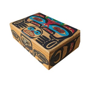 May include: A rectangular wooden box featuring a vibrant, multi-coloured graphic design on the lid and front panel. The artwork displays stylised animal motifs in black, blue, red, and green. A small metal latch is centred on the front edge.