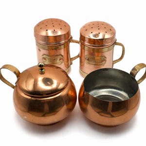 May include: A vintage copper kitchen set comprising two salt and pepper shakers, a sugar bowl with a lid, and a creamer. The set has a shiny copper finish with brass accents and handles. The salt and pepper shakers have perforated lids.