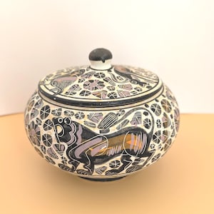 May include: A round, lidded ceramic jar with a black, white, and purple floral and animal design. The jar features a black lion illustration and a black knob on the lid. The jar is on a light orange surface.