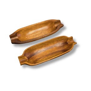 May include: Two elongated, oval-shaped wooden trays with a smooth, polished finish and visible natural wood grain patterns. Each tray features a recessed centre and small, notched indentations at both ends, showcasing a warm brown colour.