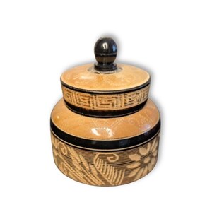 May include: A round lidded jar featuring a tan base with intricate black etched floral and leaf patterns. The lid includes a black knob handle and a geometric Greek key border design. The jar has a smooth finish with a distinct contrast between the light tan etched areas and the solid black bands that wrap around the body and the lid.
