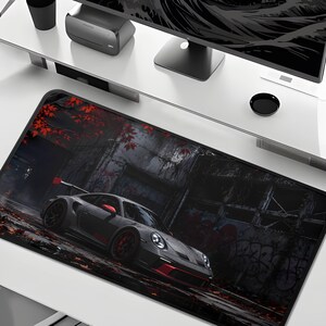 May include: A rectangular desk mat featuring a high-resolution graphic of a grey sports car with red accents parked in a dark, weathered urban setting. The scene includes fallen red autumn leaves on the ground and graffiti on the building walls.