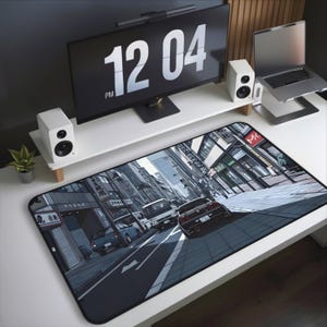 May include: A large rectangular desk mat featuring a detailed illustration of a city street scene with cars and buildings. The mat is predominantly gray and black, with white road markings. The time 12:04 PM is displayed on a monitor above.