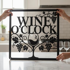 May include: Black metal sign with the words "WINE O'CLOCK" and a wine glass design. The sign also features grape clusters and vines. The sign is held up in front of a kitchen background.
