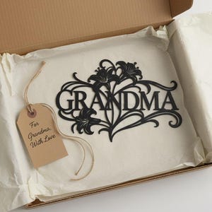 Könnte beinhalten: Ein schwarzes Metallschild mit der Aufschrift "GRANDMA" und floralen Verzierungen, in einer Pappschachtel mit Seidenpapier. Ein Geschenkanhänger mit der Aufschrift "For Grandma, With Love."
