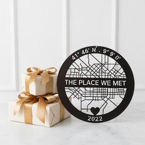 Könnte beinhalten: Ein schwarzes, kreisförmiges Wandkunstwerk mit einem Kartendesign, dem Text "THE PLACE WE MET", Koordinaten, einem Herz und dem Jahr 2022. Zwei verpackte Geschenke mit goldenen Bändern befinden sich im Hintergrund.