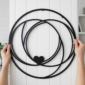 May include: Black metal wall art featuring overlapping circular shapes and a heart-shaped accent. The design is held by two hands against a white background, suggesting a decorative piece for home decor.