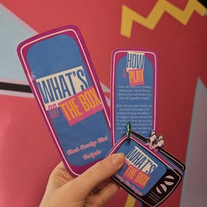 May include: A game set titled What's On The Box by Black Country Bird Budgets, featuring a tall rectangular instruction card and a stack of smaller television-shaped cards held together by a green metal ring, all with blue, pink, and yellow accents.