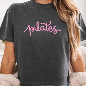 Peut inclure: Un t-shirt à manches courtes gris anthracite avec le mot pilates écrit en police rose cursive et agrémenté de petites lignes vertes. Le vêtement présente une encolure ronde, une coupe décontractée et une texture de tissu délavée.