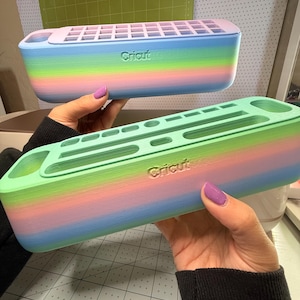 May include: Two rainbow-colored Cricut pen holders are held up. The holders are rectangular with a grid on top and the Cricut logo on the side. The colors transition from blue to pink to green.