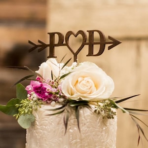 Könnte beinhalten: Eine weiße Hochzeitstorte mit einem Holz-Topper, auf dem "P&D" mit einem Pfeil durch ein Herz steht. Die Torte ist mit weißen Rosen und rosa Blumen verziert.