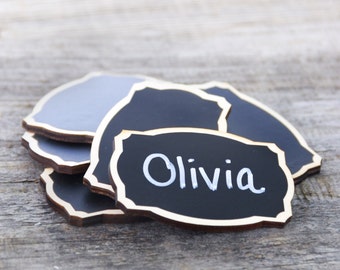 May include: A stack of black chalkboard place cards with a gold border. The top card has the name Olivia written in white chalk.
