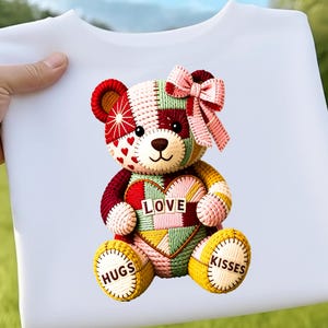 May include: White t-shirt with a colourful teddy bear design. The bear has a patchwork design with a heart that says "LOVE". The bear also has a pink bow, and the feet say "HUGS" and "KISSES".