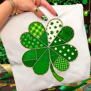 Può includere: Cuscino bianco con un grande trifoglio applicato. Il trifoglio è composto da vari tessuti verdi fantasia, tra cui pois, quadri e stampe floreali, con bordatura dorata. Una mano con un anello e un braccialetto d'oro tiene il cuscino.