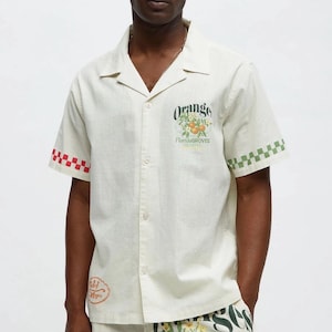 Puede incluir: Camisa crema con botones y pantalones cortos a juego con gráficos de naranjas y flores. La camisa presenta el texto "Orange Florida Groves" y puños a cuadros. Los pantalones cortos también tienen el texto "Florida Groves" y gráficos de naranjas.